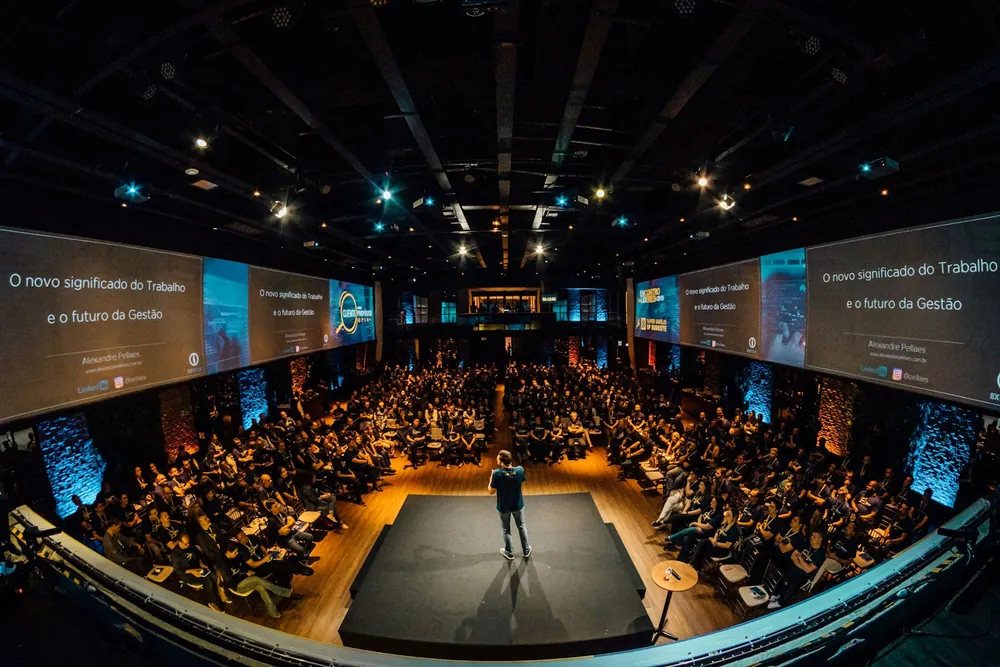 Auditorio lleno con un orador en el escenario y pantallas laterales